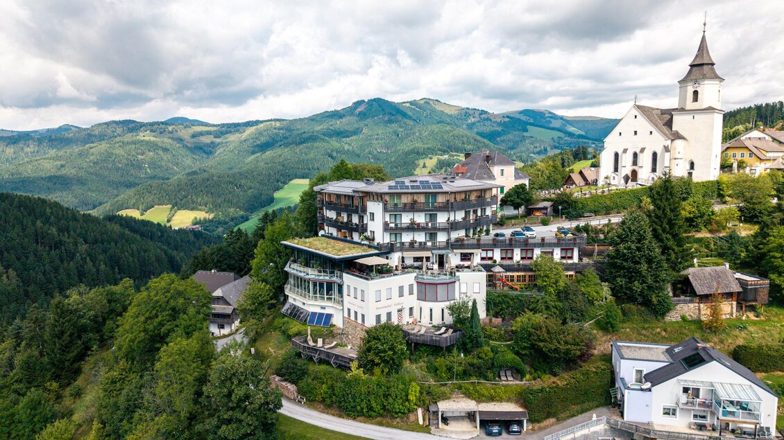 Ein malerisches Hotelgebäude in einer bergigen Umgebung. Im Hintergrund sind sanfte Hügel und eine Kirche sichtbar. | © Oststeiermark Tourismus