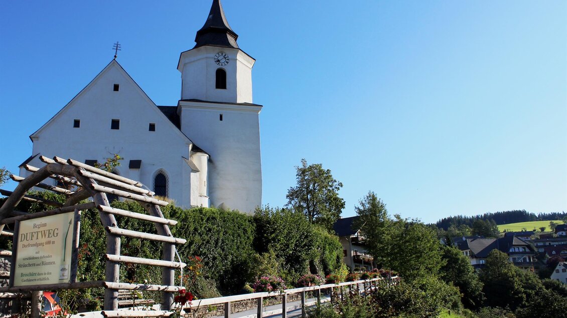 Eine charmante Kirche mit einem spitzen Turm, umgeben von Bäumen und einem klaren blauen Himmel. Im Vordergrund befindet sich ein schöner Garten mit einem Holzpfad. | © Tourismusverband Oststeiermark