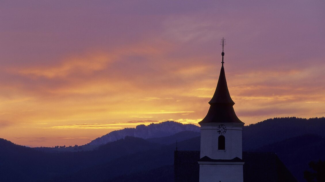 Eine Kirche mit einem spitzen Turm steht vor einem malerischen Sonnenuntergang. Die Silhouette der Berge ergänzt die idyllische Szene. | © Tourismusverband Oststeiermark