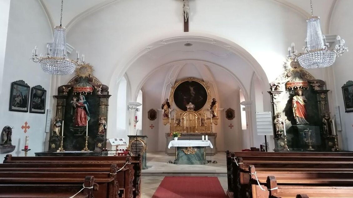 Ein heller Kircheninnenraum mit Holzbänken und einem Altar. An den Seiten stehen prächtig verzierte Statuen und ein Kruzifix hängt an der Decke. | © TVB Ausseerland Salzkammergut/Kolb