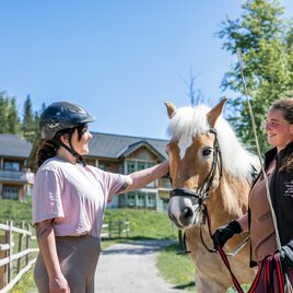 Two women are standing outdoors and interacting with a horse. Buildings and trees can be seen in the background. | © www.zloam.at