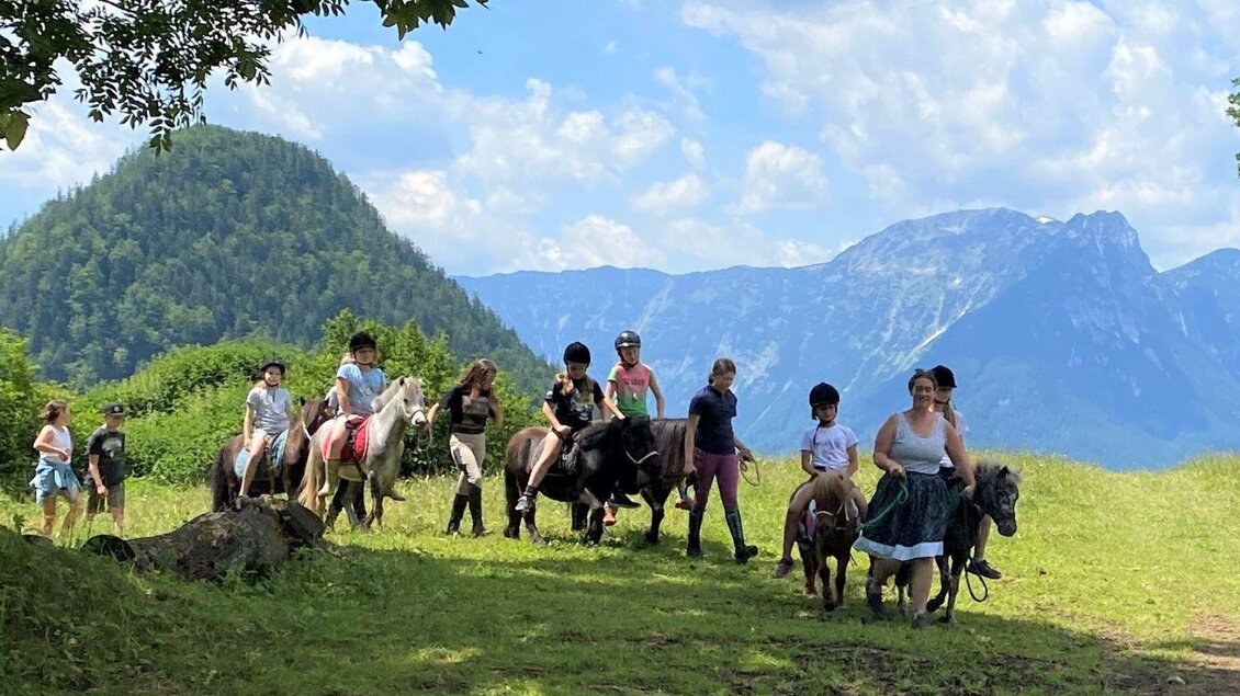 Eine Gruppe von Kindern reitet auf Ponys in einer grünen Landschaft. Im Hintergrund sind Berge und ein blauer Himmel mit einigen Wolken zu sehen. | © Pferdefreunde Zloam, www.pferdefreunde-zloam.at