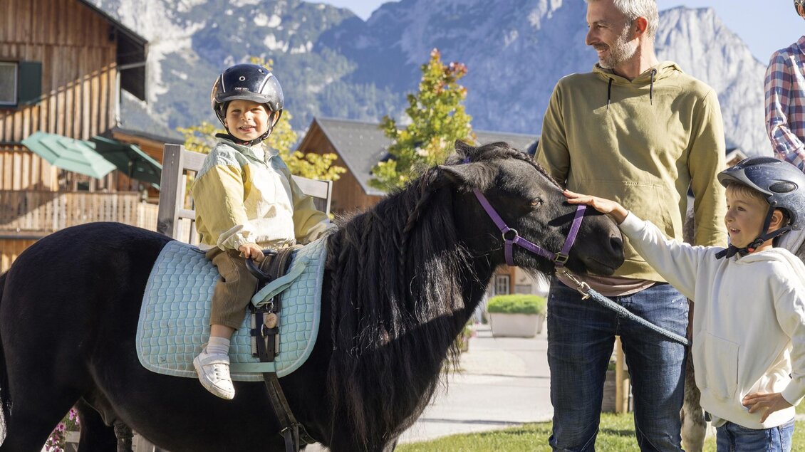 Ein Kind sitzt auf einem kleinen Pferd, während ein Erwachsener und ein anderes Kind daneben stehen. Im Hintergrund sind Berge und eine ländliche Umgebung zu sehen. | © www.zloam.at