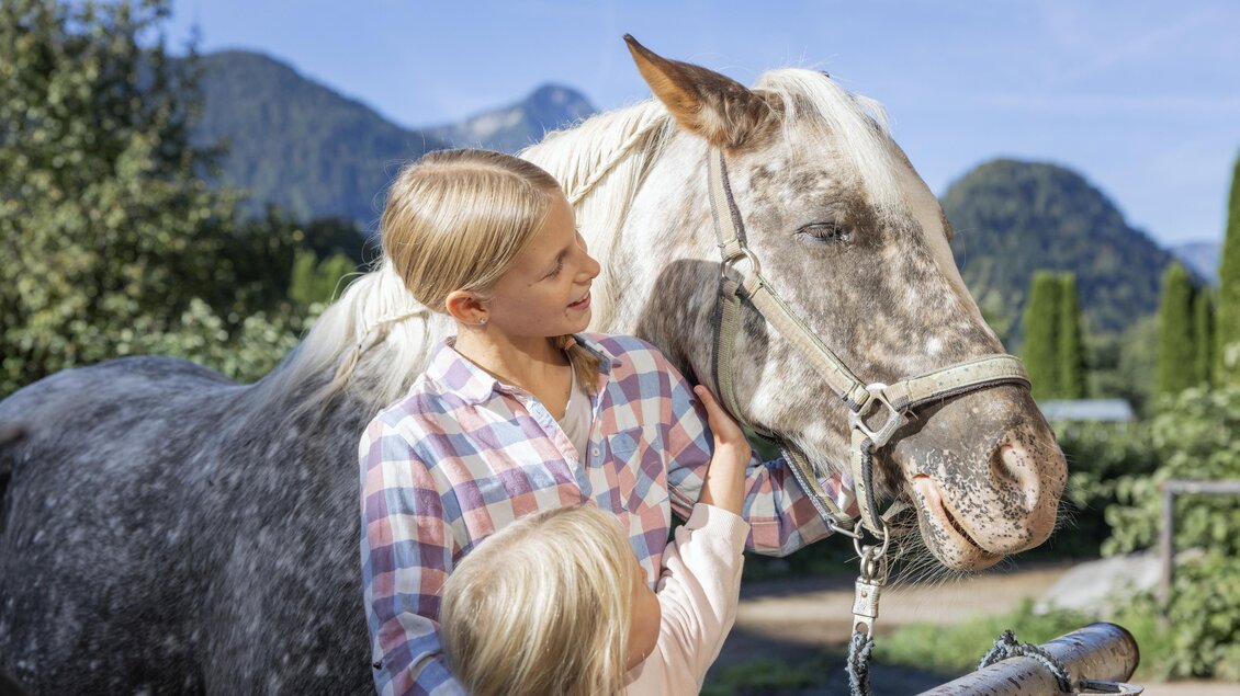Ein Mädchen streichelt ein Pferd in einer ländlichen Umgebung. Im Hintergrund sind Bäume und Berge zu sehen. | © www.zloam.at