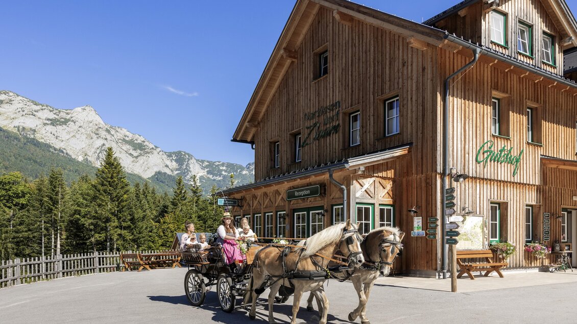 Eine Holzfarbe mit einem traditionellen Gebäude und einer Kutsche, die von zwei Pferden gezogen wird. Im Hintergrund sind Berge und grüne Bäume zu sehen. | © www.zloam.at