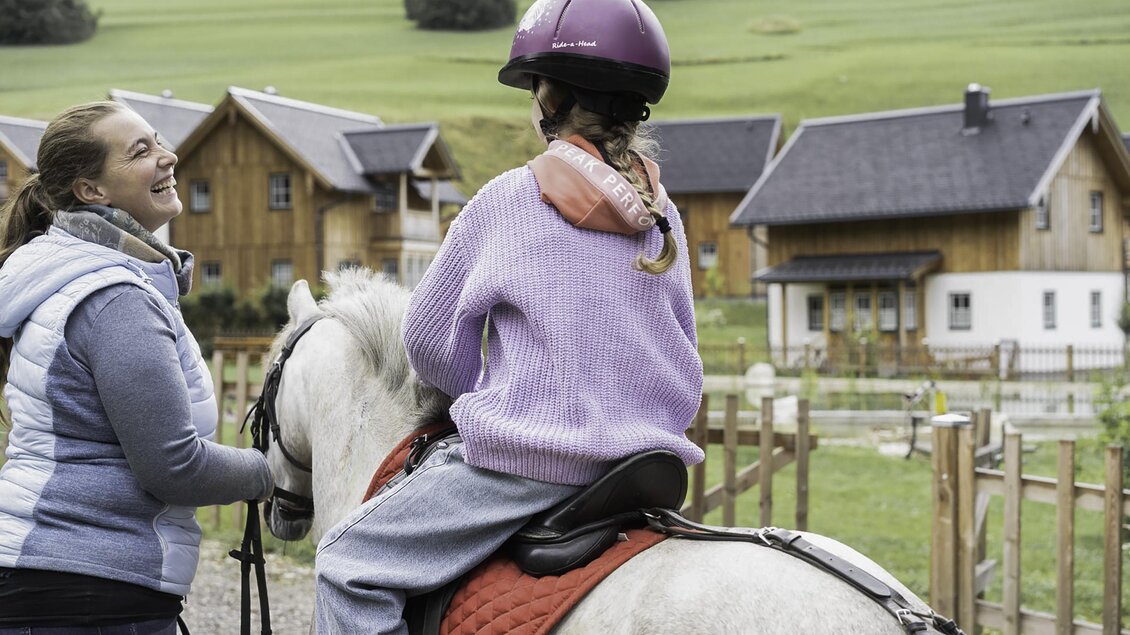 Eine Frau spricht lächelnd mit einem Mädchen auf einem Pferd. Im Hintergrund sind Holzhäuser und Wiesen zu sehen. | © Pferdefreunde Zloam, www.pferdefreunde-zloam.at