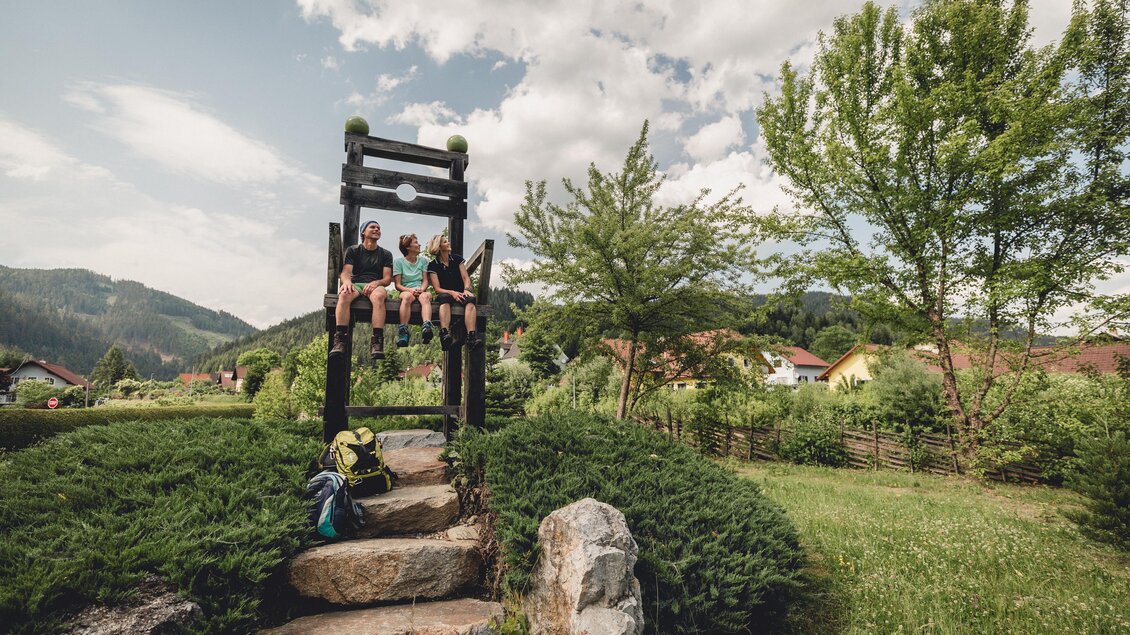Zwei Personen sitzen auf einer Holzbank in einer grünen Gartenlandschaft. Im Hintergrund sind Bäume und Hügel sichtbar. | © TV Hochsteiermark, nicoleseiser.at