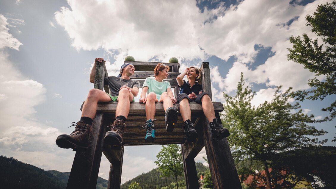 Drei Personen sitzen auf einer hohen Holzbank im Freien. Der Himmel ist bewölkt und es gibt viel grüne Natur um sie herum. | © TV Hochsteiermark, nicoleseiser.at