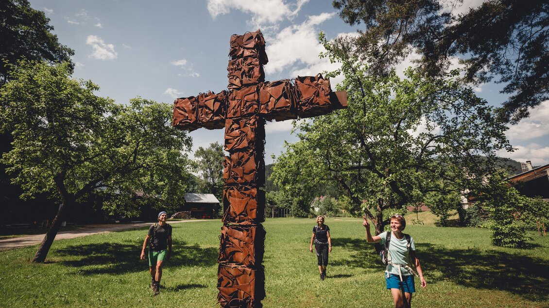 Drei Personen gehen in einer grünen Landschaft. Im Vordergrund steht ein kunstvoll gestaltetes Holzkreuz. | © TV Hochsteiermark, nicoleseiser.at