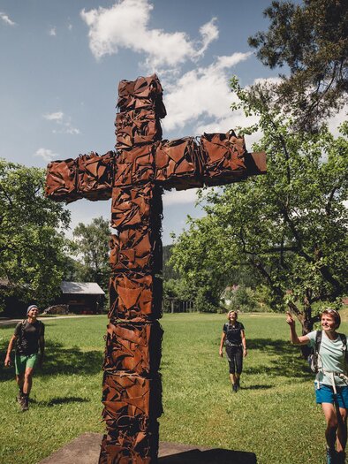 Drei Personen gehen in einer grünen Landschaft. Im Vordergrund steht ein kunstvoll gestaltetes Holzkreuz. | © TV Hochsteiermark, nicoleseiser.at