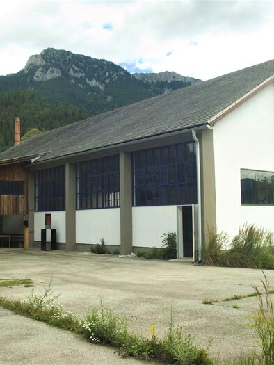 A modern building with large windows and a wooden facade stands against a mountain backdrop. In the foreground, a sculpture can be seen, surrounded by green spaces. | © Susanna Tabaka Pillhofer