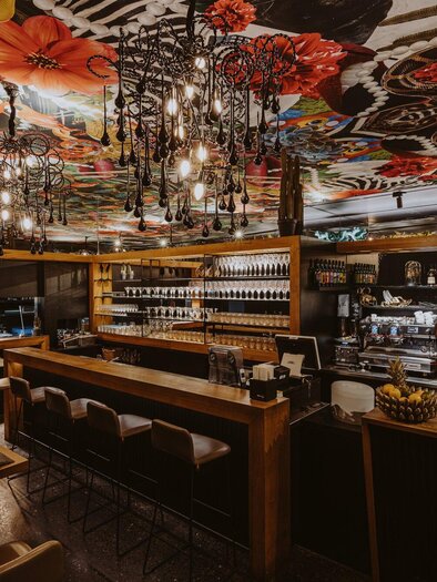 Ein stilvolles Restaurant mit einer langen Bar und eleganter Beleuchtung. Die Decke ist mit bunten Blumenmustern dekoriert und schafft eine einladende Atmosphäre. | © Aiola - Joel Kernasenko