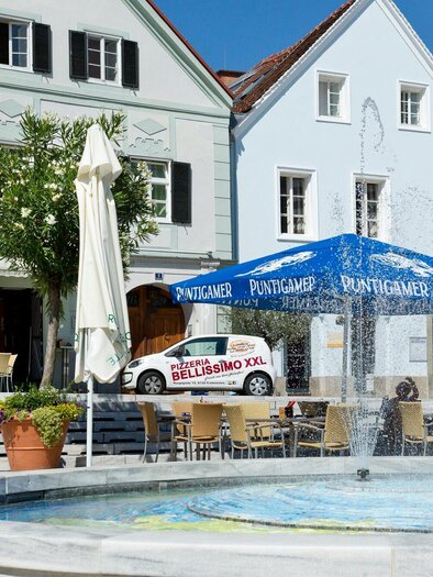 A beautiful square with a fountain and colorful buildings. There are chairs and tables under a parasol. | © TV Region Graz - René Vidalli