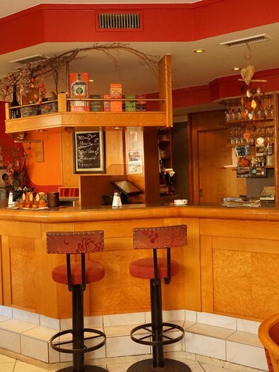 Eine gemütliche Bar mit einer warmen orangefarbenen Wandgestaltung. Auf der Theke stehen Glasware und Dekoration, und es gibt Sitzgelegenheiten für Gäste. | © Pizzeria Draxler