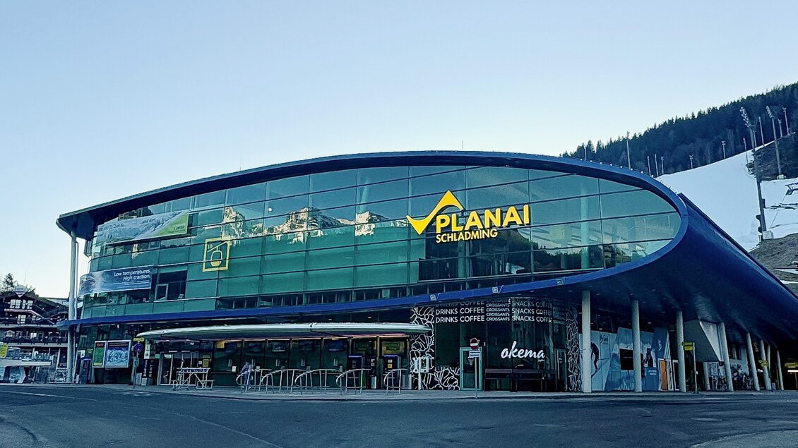 Ein modernes Gebäude mit großen Glasfenstern, das als Ticket- und Informationszentrum für die Planai in Schladming dient. Draußen warten mehrere Menschen auf den Ski-Service. | © Sabrina Melcher, Planai