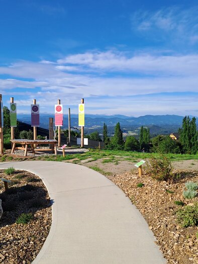 A beautiful garden with a path leading to colorful flags. In the background, hills and a wine barrel can be seen. | © TV Südsteiermark