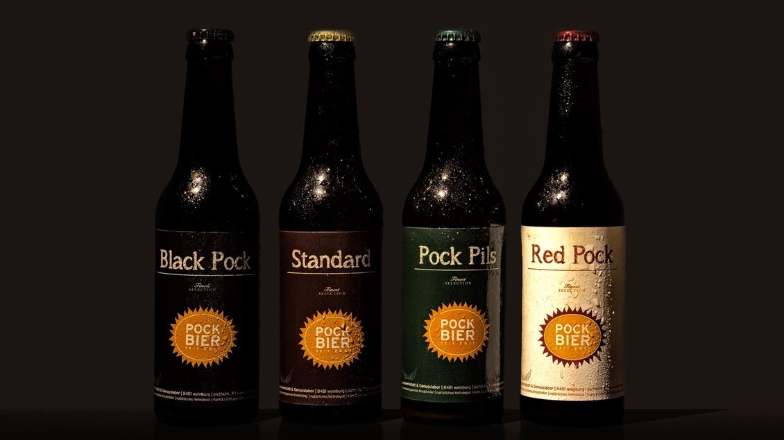 Vier Bierflaschen stehen vor einem dunklen Hintergrund. Die Etiketten zeigen verschiedene Sorten: Black Pock, Standard, Pock Pils und Red Pock. | © GlaMur
