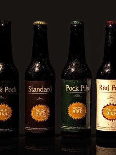 Vier Bierflaschen stehen vor einem dunklen Hintergrund. Die Etiketten zeigen verschiedene Sorten: Black Pock, Standard, Pock Pils und Red Pock. | © GlaMur