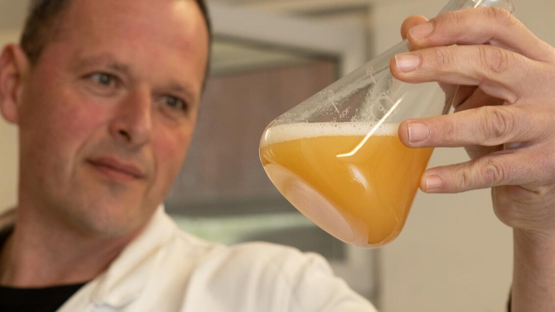 Ein Wissenschaftler in einem weißen Laborkittel betrachtet eine Erlenmeyerkolben mit orangefarbigem Inhalt. Der Forscher analysiert sorgfältig die Flüssigkeit. | © GlaMur