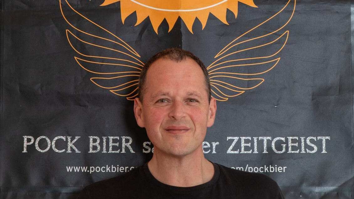 Ein Mann steht vor einem Banner mit dem Schriftzug "POCK BIER". Er trägt ein schwarzes T-Shirt mit dem Aufdruck "FIRST MATE". | © GlaMur