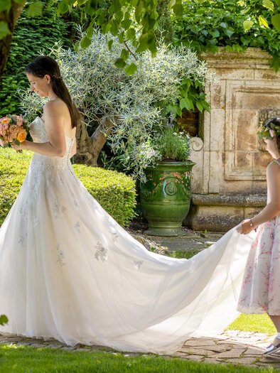 Eine Braut in einem eleganten weißen Kleid geht durch einen schönen Garten. Ein kleines Mädchen hält das Ende des Kleides und schaut zu ihr auf. | © Renate Polz