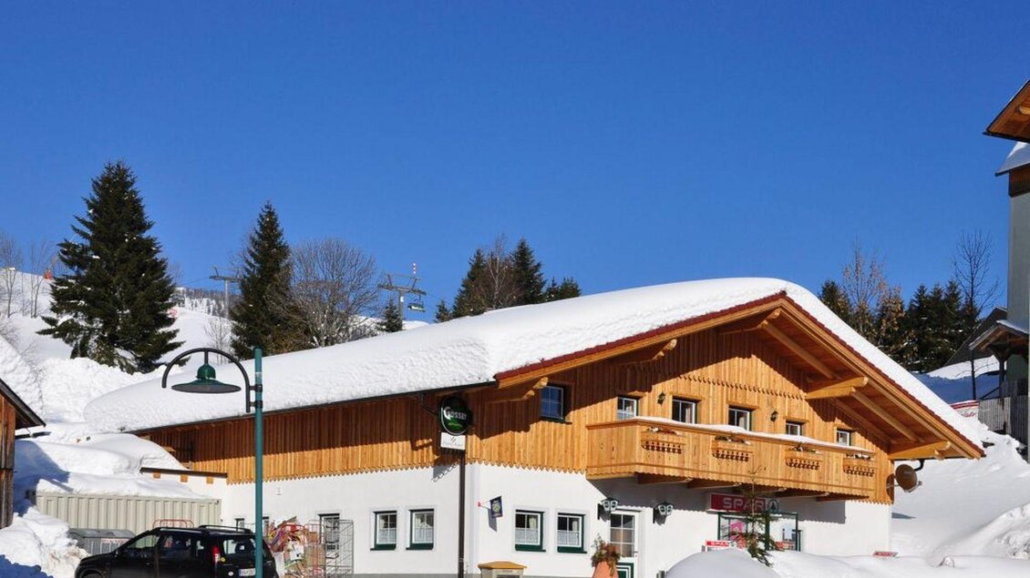 Ein gemütliches Holzhaus im Schnee unter einem klaren blauen Himmel. Umgeben von Tannenbäumen und winterlicher Landschaft. | © Familie Heiß