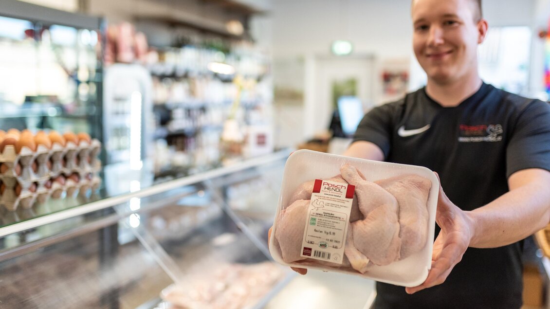 Ein Verkäufer präsentiert ein Paket mit Hähnchenfleisch in einem Lebensmittelgeschäft. Im Hintergrund sind Regale mit weiteren Lebensmitteln zu sehen. | © RM SW GmbH | webquartier.at
