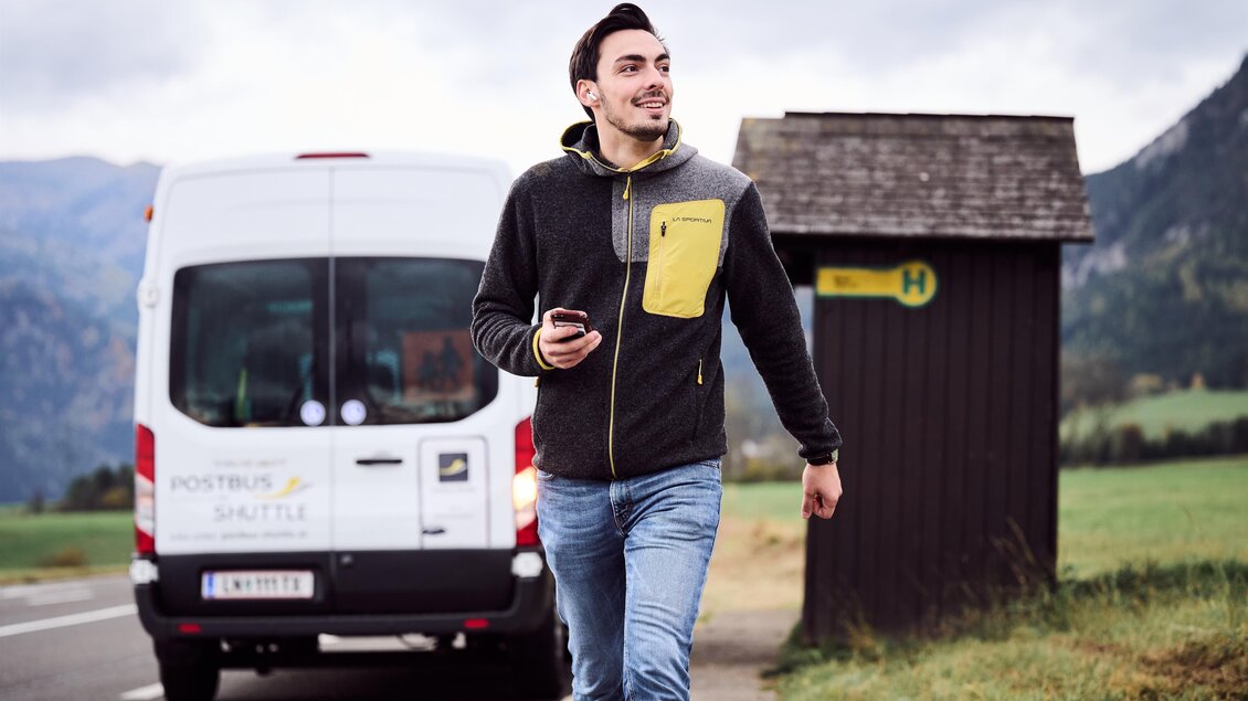Ein junger Mann geht lächelnd auf einer Landstraße, während er sein Handy in der Hand hält. Im Hintergrund ist ein Bus und eine Haltestelle sichtbar. | © ÖBB/ Marek Knopp