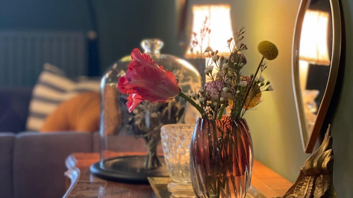 Ein stilvoller Tisch mit einer Vase voller Blumen und dekorativen Gegenständen. Im Hintergrund sind sanfte Lichter und eine gemütliche Einrichtung zu sehen. | © Principessa 1st bar