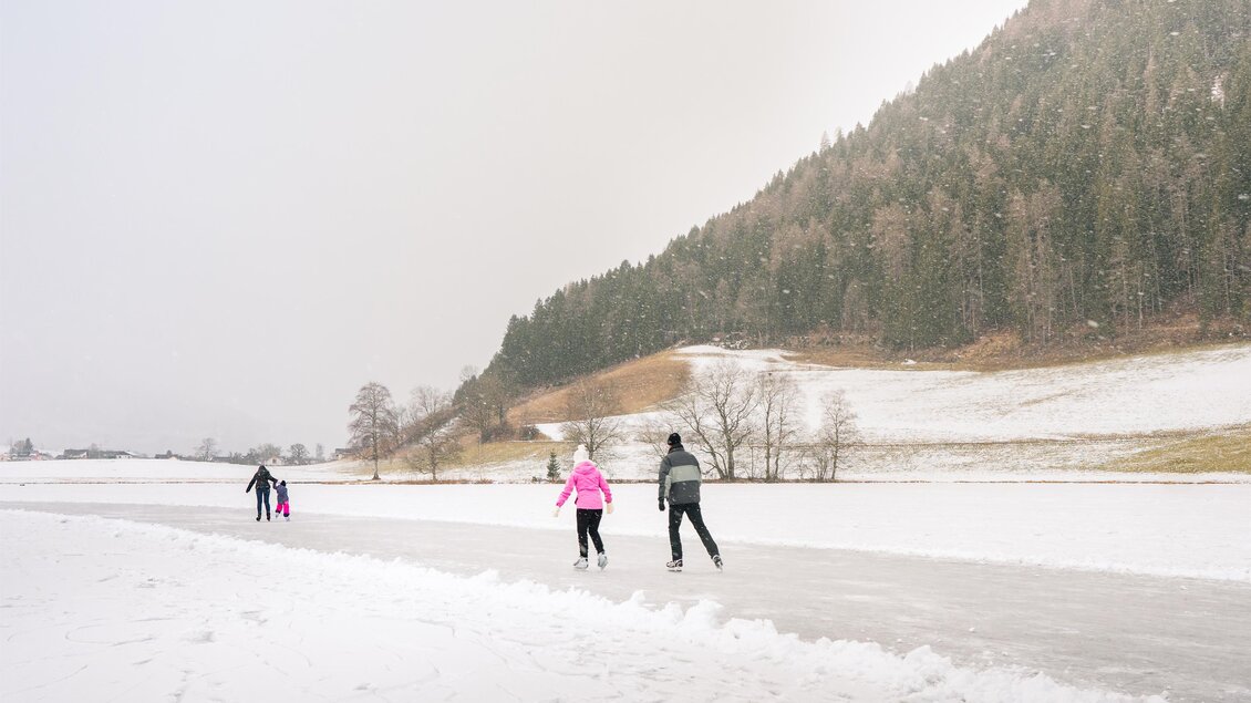 Eine winterliche Landschaft mit einer zugefrorenen Fläche, auf der mehrere Personen Schlittschuh laufen. Der Schnee bedeckt die Umgebung und die Bäume im Hintergrund sind grün. | © Christoph Lukas