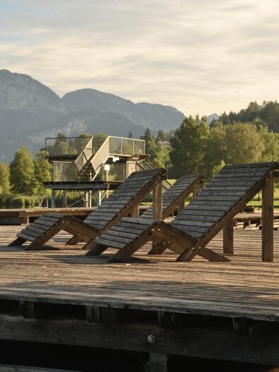 A wooden terrace by the lake with loungers. In the background, mountains and trees can be seen. | © Putterersee