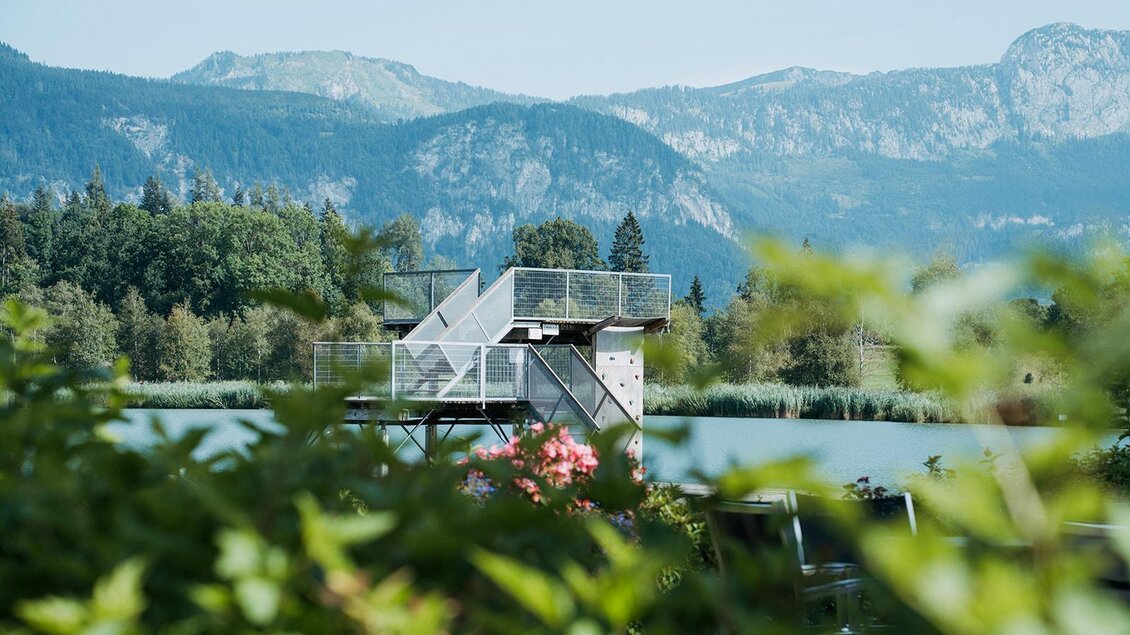 Ein Aussichtsturm am Ufer eines ruhigen Sees, umgeben von grünen Bäumen. Im Hintergrund sind beeindruckende Berge zu sehen.