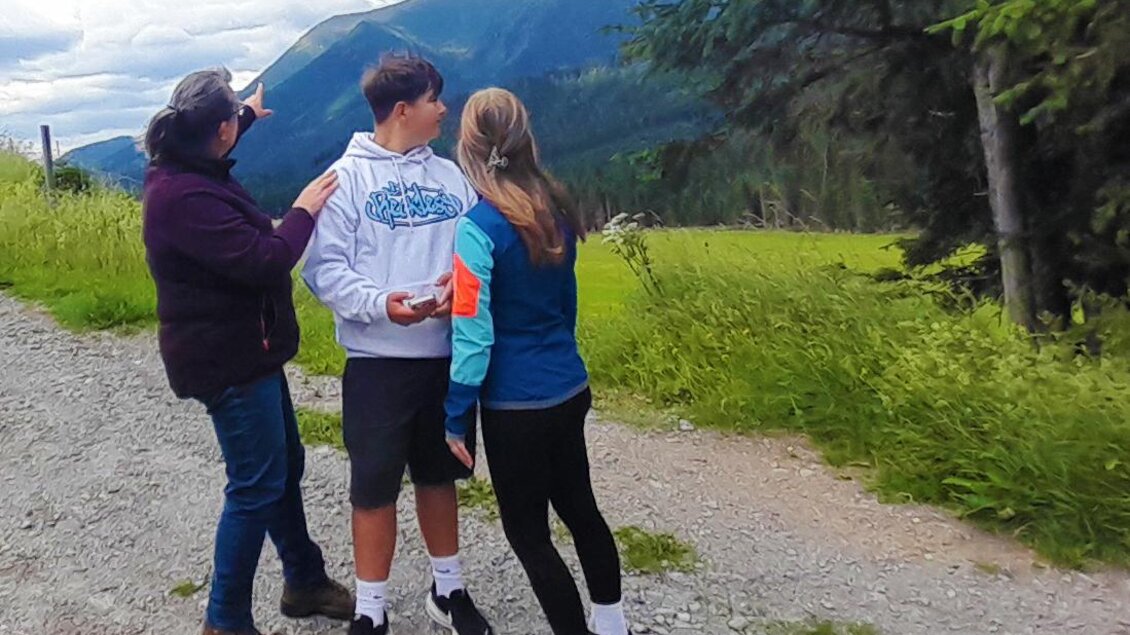 Drei Personen stehen auf einem Schotterweg und schauen in die Ferne. Im Hintergrund sind grüne Wiesen und Berge zu sehen. | © Loes Dikmans