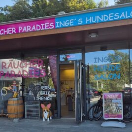 Eine Haustier-Geschäftsfront mit dem Schild „Räucher Paradies“ und „Inge's Hundesalon“. Vor der Tür steht ein Schaufenster mit einer Hundeabbildung und einem Fahrrad. | © Räucher Paradies