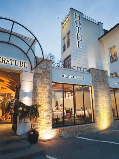 Das Bild zeigt den Eingang eines Hotels mit dem Schriftzug "Römerstube". Die Fassade ist beleuchtet und hat eine moderne Architektur. | © Römerstube