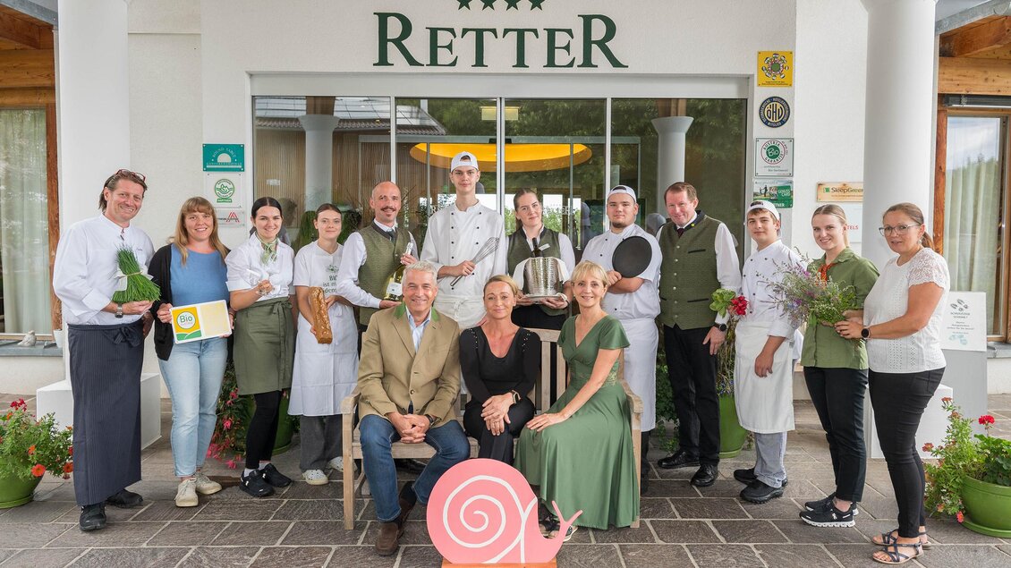Eine Gruppe von Mitarbeitern vor einem Restaurant mit dem Namen "Retter". Sie halten verschiedene Utensilien und Pflanzen, während sie lächeln und in die Kamera schauen. | © RETTER Bio-Natur-Resort, Bio Austria, Slow Food Österreich, Helmut Schweighofer