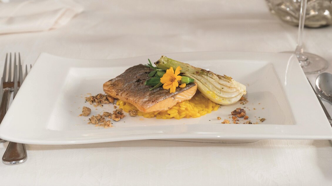 Ein elegantes Gericht mit gebratenem Fisch auf einem Bett aus Risotto. Das Essen ist mit frischen Kräutern und einer Blume dekoriert. | © RETTER Bio-Natur-Resort, Helmut Schweighofer