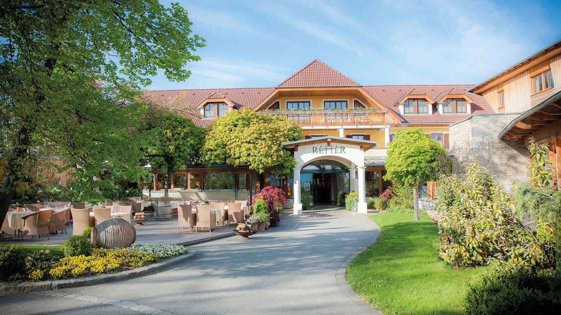 Ein schönes Hotel mit einem einladenden Eingang und umgeben von Bäumen und Blumen. Auf der Terrasse stehen Tische und Stühle, ideal für Gäste im Freien. | © RETTER Bio-Natur-Resort