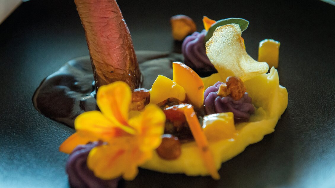 Ein kunstvoll angerichteter Teller mit zartem Fleisch, bunten Beilagen und essbaren Blumen. Die Komposition wirkt elegant und ansprechend. | © RETTER Bio-Natur-Resort, Helmut Schweighofer