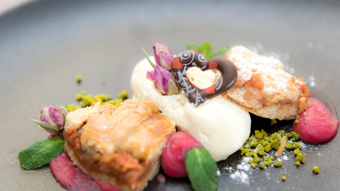 Ein kunstvoll angerichtetes Dessert auf einem schwarzen Teller. Es besteht aus einer cremigen Basis, umgeben von bunten Früchten, essbaren Blüten und einer dekorativen Schokoladendekoration. | © RETTER Bio-Natur-Resort, Helmut Schweighofer