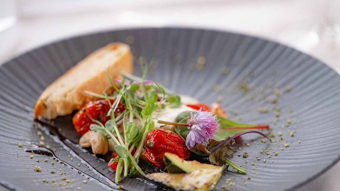 Ein stilvoll angerichteter Teller mit gerösteten Tomaten, frischem Gemüse und einem Stück Brot. Das Gericht wird von einer feinen Sauce und essbaren Blumen garniert. | © RETTER Bio-Natur-Resort, Helmut Schweighofer