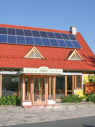 A modern house with a red roof and solar panels. It has a yellow facade and is surrounded by green plants. | © Helmut Schweighofer