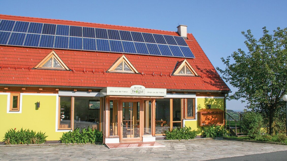 Ein gelbes Haus mit einem roten Dach und Solarpanelen. Die Fassade hat große Fenster und ist von Grünanlagen umgeben. | © RETTER BioGut