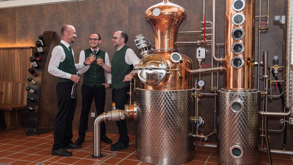Drei Männer in traditioneller Kleidung stehen neben einer großen Kupfer-Destille. Die Atmosphäre ist freundlich und rustikal, mit einer modernen Brennanlage im Hintergrund. | © RETTER BioGut