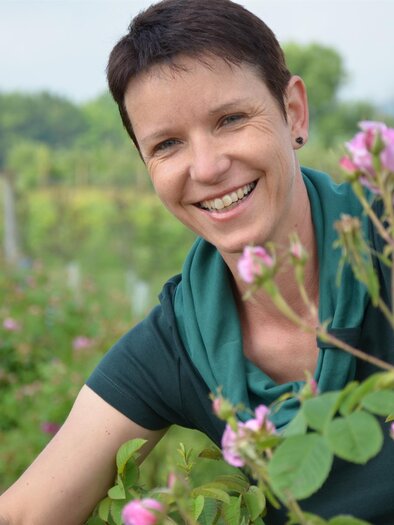 Eine lächelnde Frau steht zwischen Rosensträuchern. Sie genießt die Natur und die blühenden Blumen im Hintergrund. | © Raabauer Eisvogel