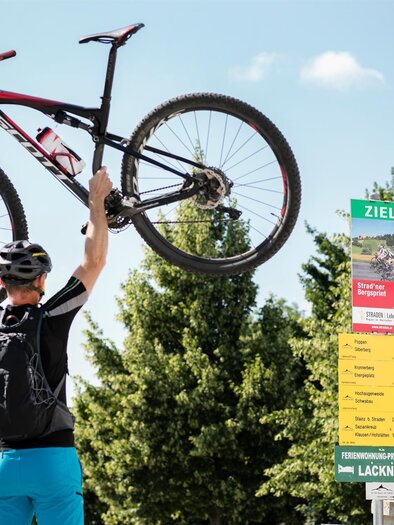 Ein Radfahrer hebt sein Mountainbike über den Kopf. Im Hintergrund ist ein Wegweiser mit dem Ziel angegeben. | © Harald Eisenberger