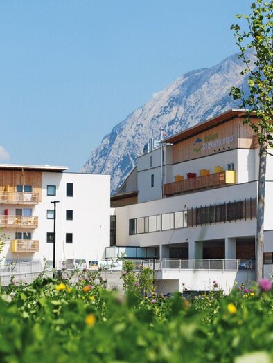 <p>Das Moderne Aldiana Club Hotel mit Holzakzenten, umgeben von grünen Wiesen und blühenden Blumen. Im Hintergrund ist der beeindruckende Grimming sichtbar.</p> | © Karin Ackermann