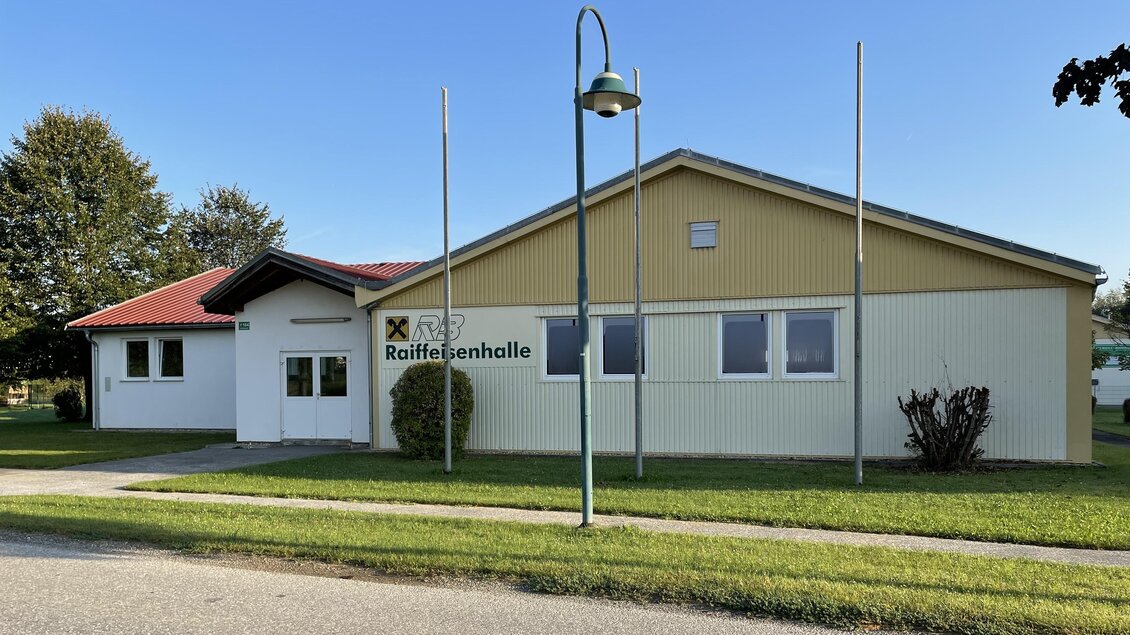 <p>Ein modernes Gebäude mit einem flachen Dach und hellen Fassaden. Vor dem Gebäude befinden sich Rasenflächen und einige Bäume. Raiffeisenhalle für Veranstaltungen in Buch bei Geiseldorf.</p>