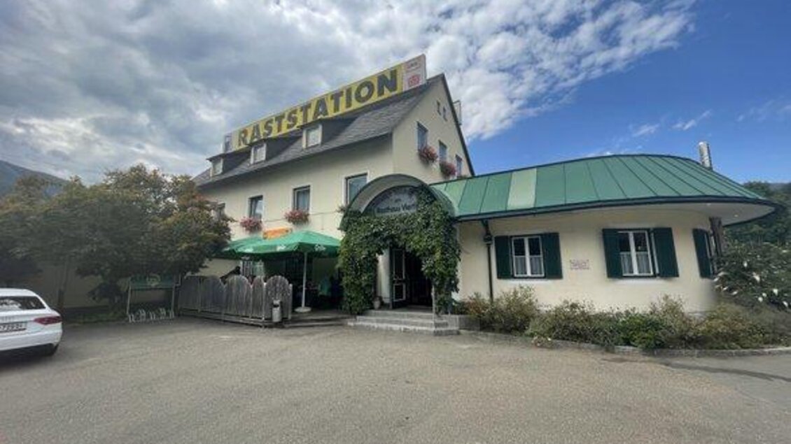 Ein Restaurant mit dem Namen "Raststation" in einer ländlichen Umgebung. Das Gebäude hat eine grüne Dachsäule und ist von Bäumen und Pflanzen umgeben. | © Klemens Draxl