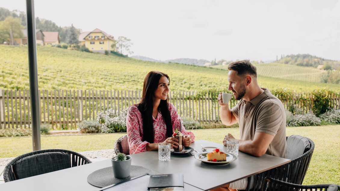 Ein Paar sitzt an einem Tisch im Freien und genießt ein Getränk. Im Hintergrund sind Weinberge und ein Haus zu sehen. | © Ratscher Landhaus
